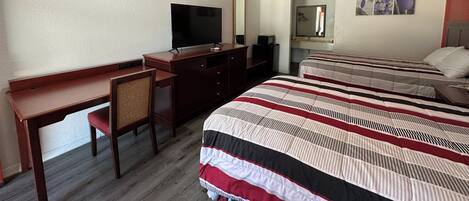 A hotel room with a desk, a television, a bed with striped bedding, a mirror, and a small artwork on the wall.