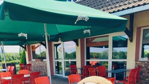 Un gran personaje de osito Teddy sentado en una silla en el patio de un restaurante, con sillas rojas y sombrillas verdes.