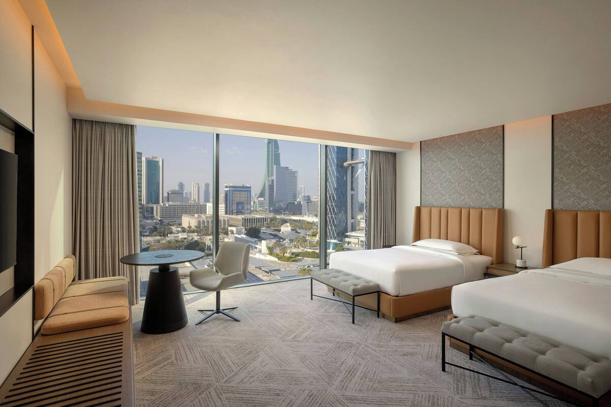 A hotel room with two beds, a sofa, a small table, and a large window offering a city view.