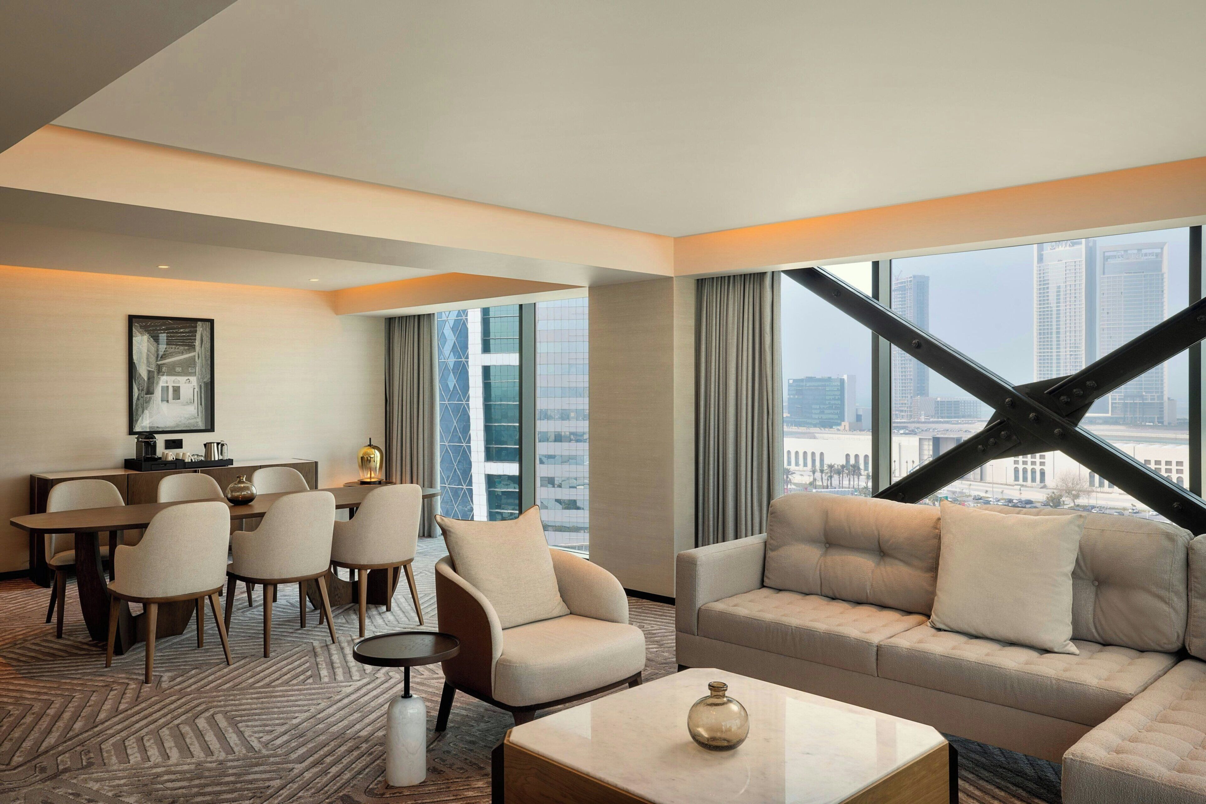 A modern hotel room with a dining area, a sofa, and a large window with a city view.