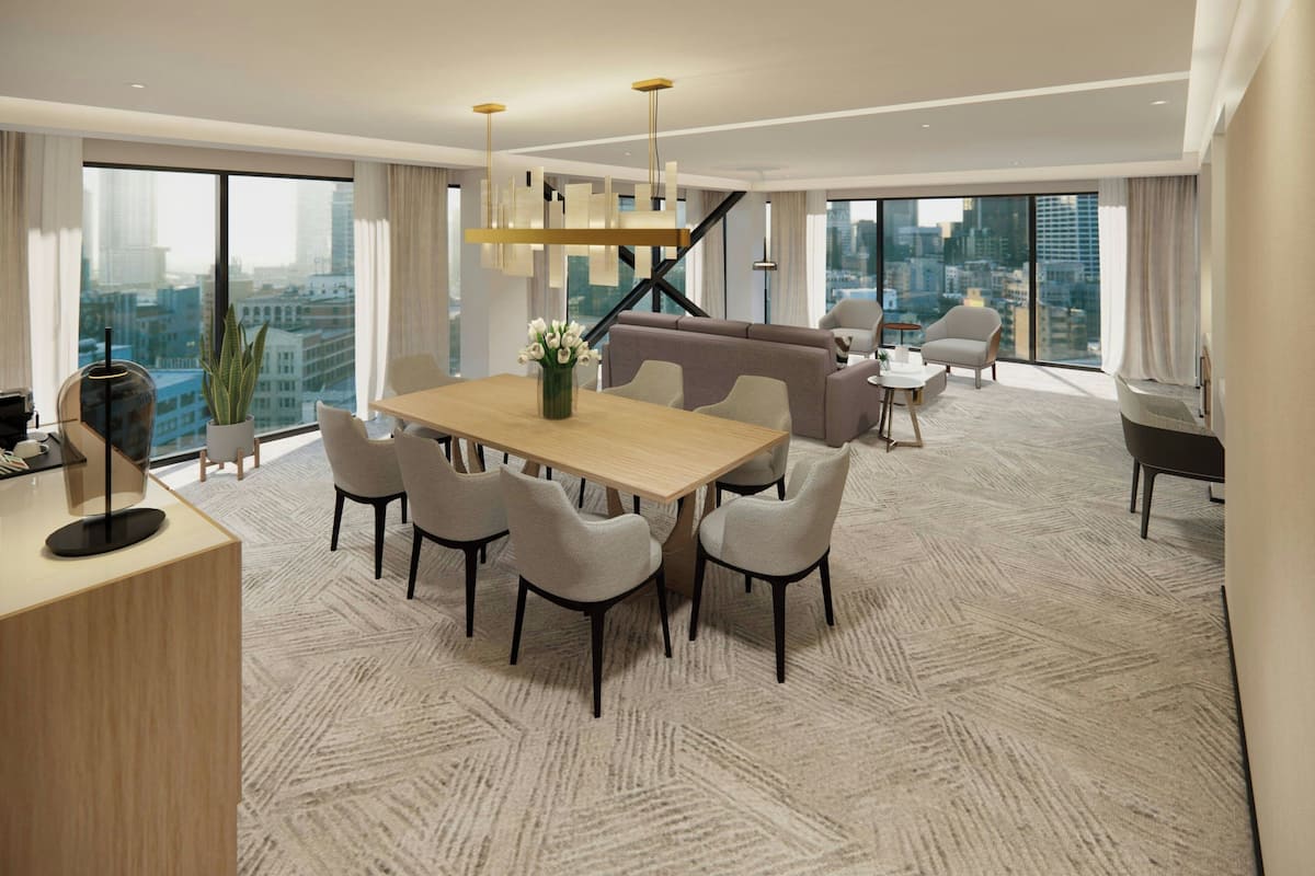 A modern hotel room with a large dining table, comfortable seating, a city view, and a spacious layout.