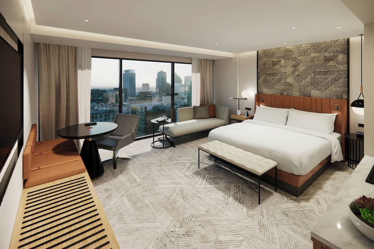 A modern hotel room with a large bed, a sofa, a dining table, and a city view.