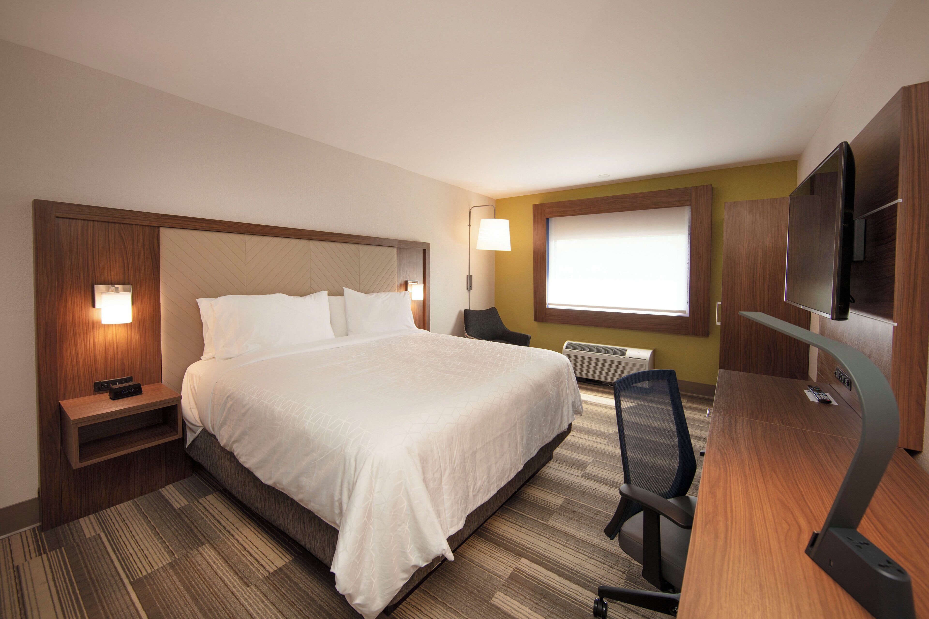 Une chambre d’hôtel avec un grand lit, des tables de chevet, un bureau avec une chaise, une télévision et une fenêtre avec des stores.