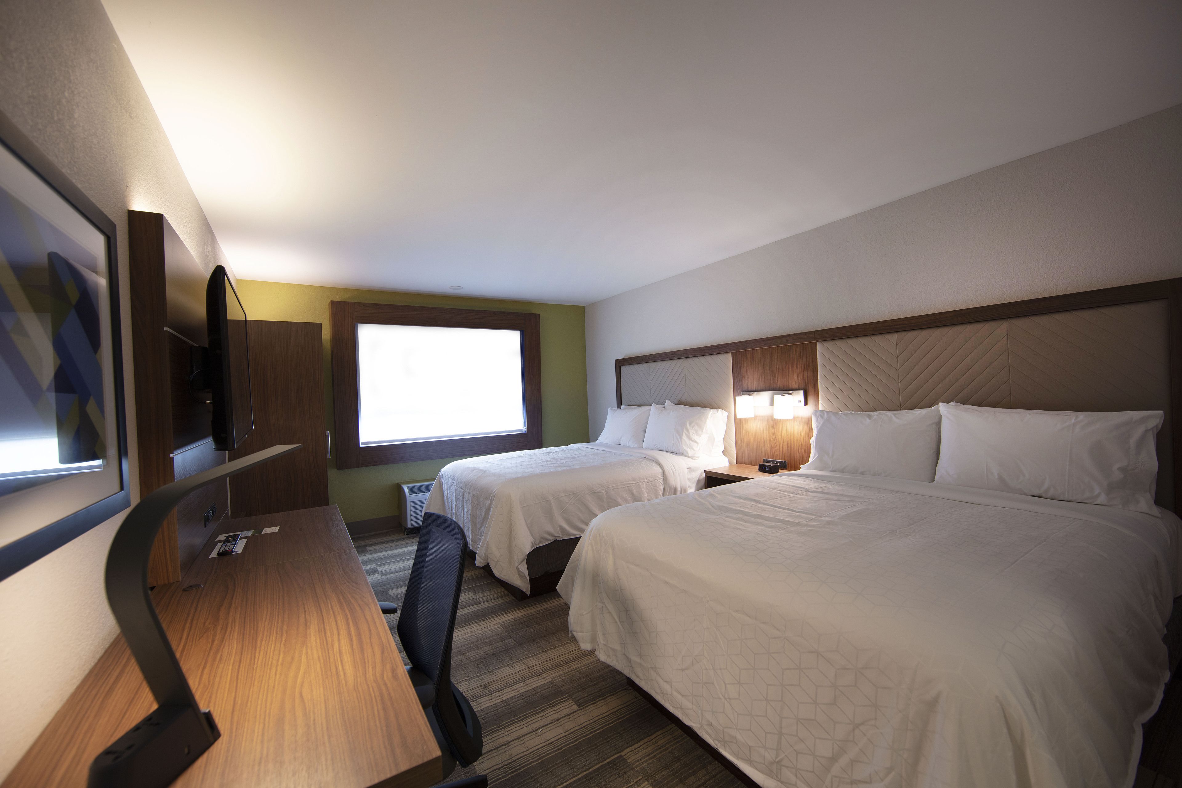 A hotel room with two beds, a desk, a chair, a TV, and a window providing natural light.