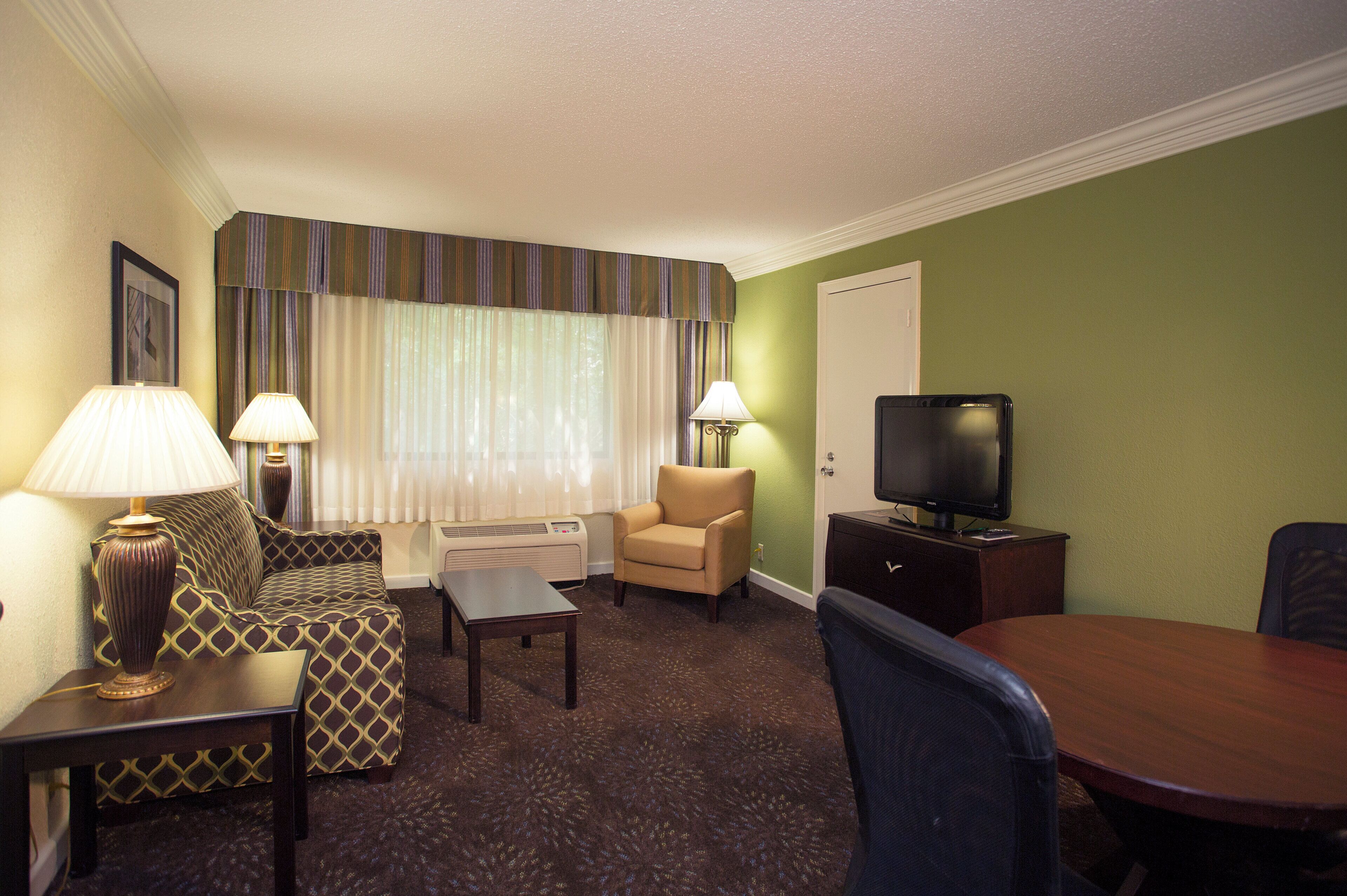 A hotel room with a sofa, armchair, TV, lamp, and a dining table.