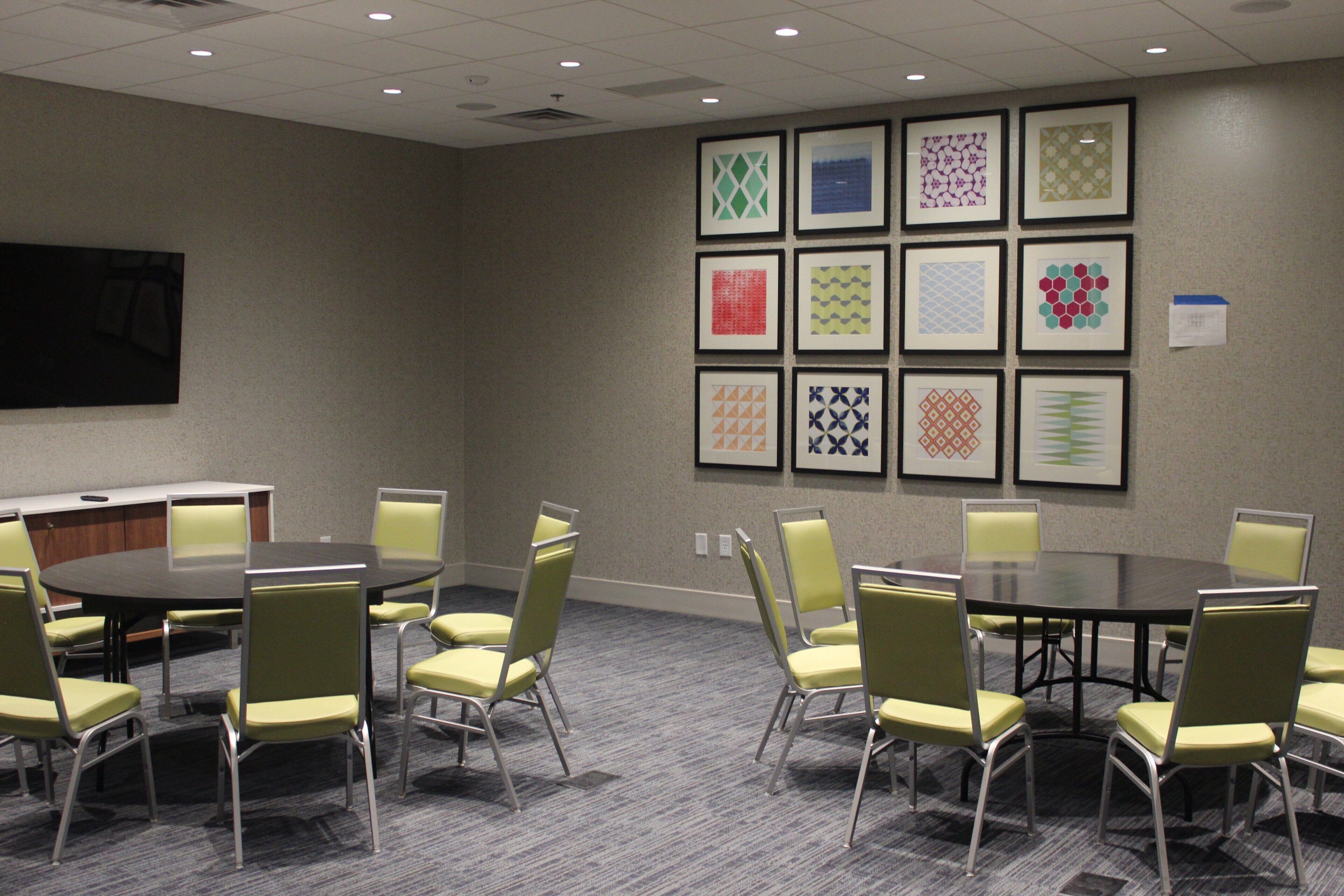 A conference room with a round table and chairs, a projector screen, and a wall with framed geometric patterns.