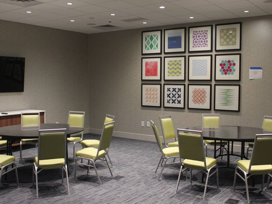 Un salón de conferencias con una mesa redonda y sillas, una pantalla de proyector y una pared con motivos geométricos enmarcados.