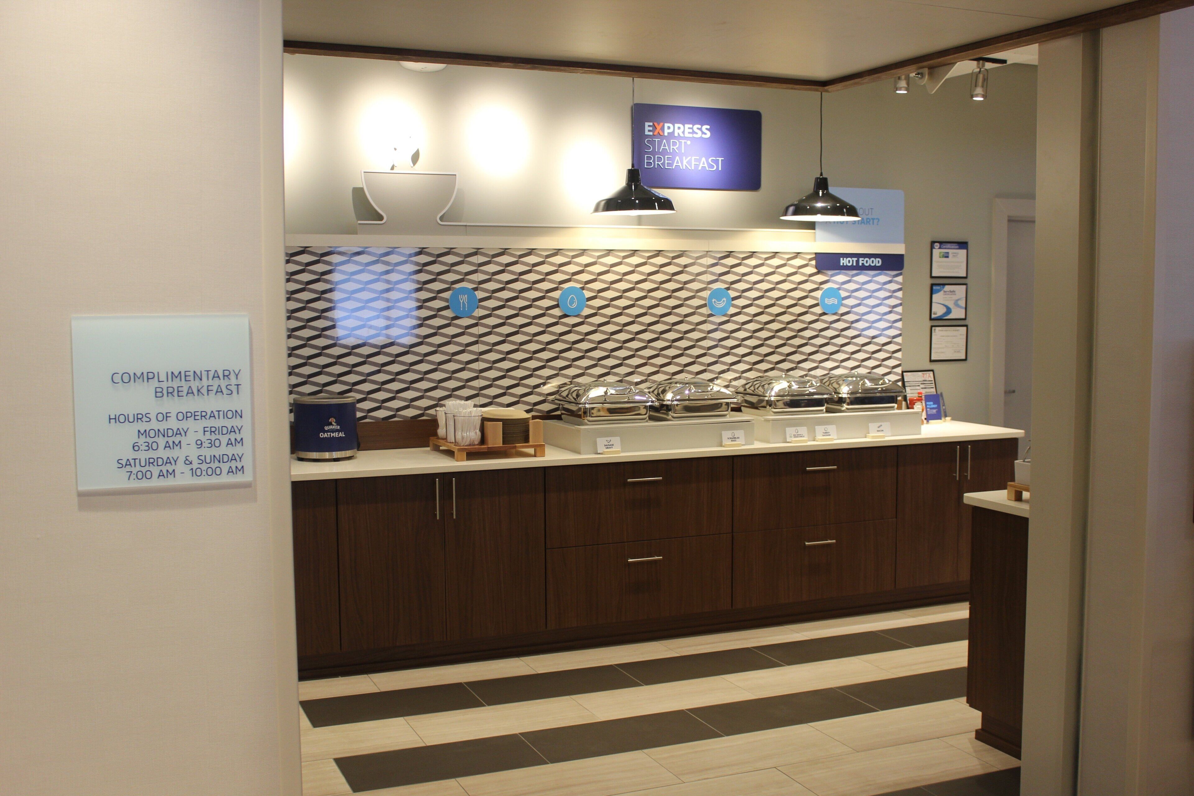 A modern kitchen area with a sign indicating complimentary breakfast hours, a countertop with various kitchen appliances, and a patterned backsplash.
