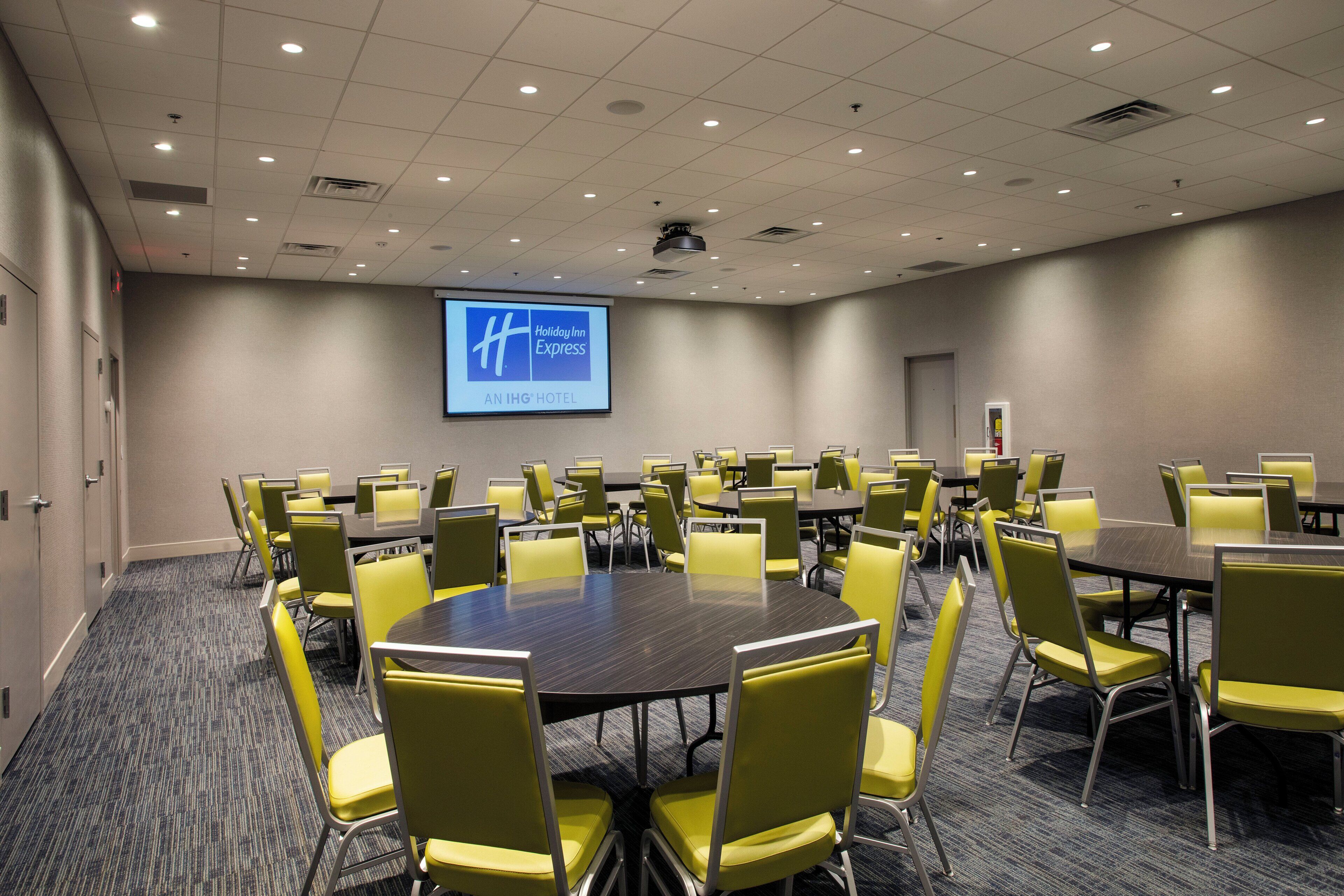 A conference room with multiple tables and chairs, a large screen displaying the Holiday Inn Express logo, and a carpeted floor.