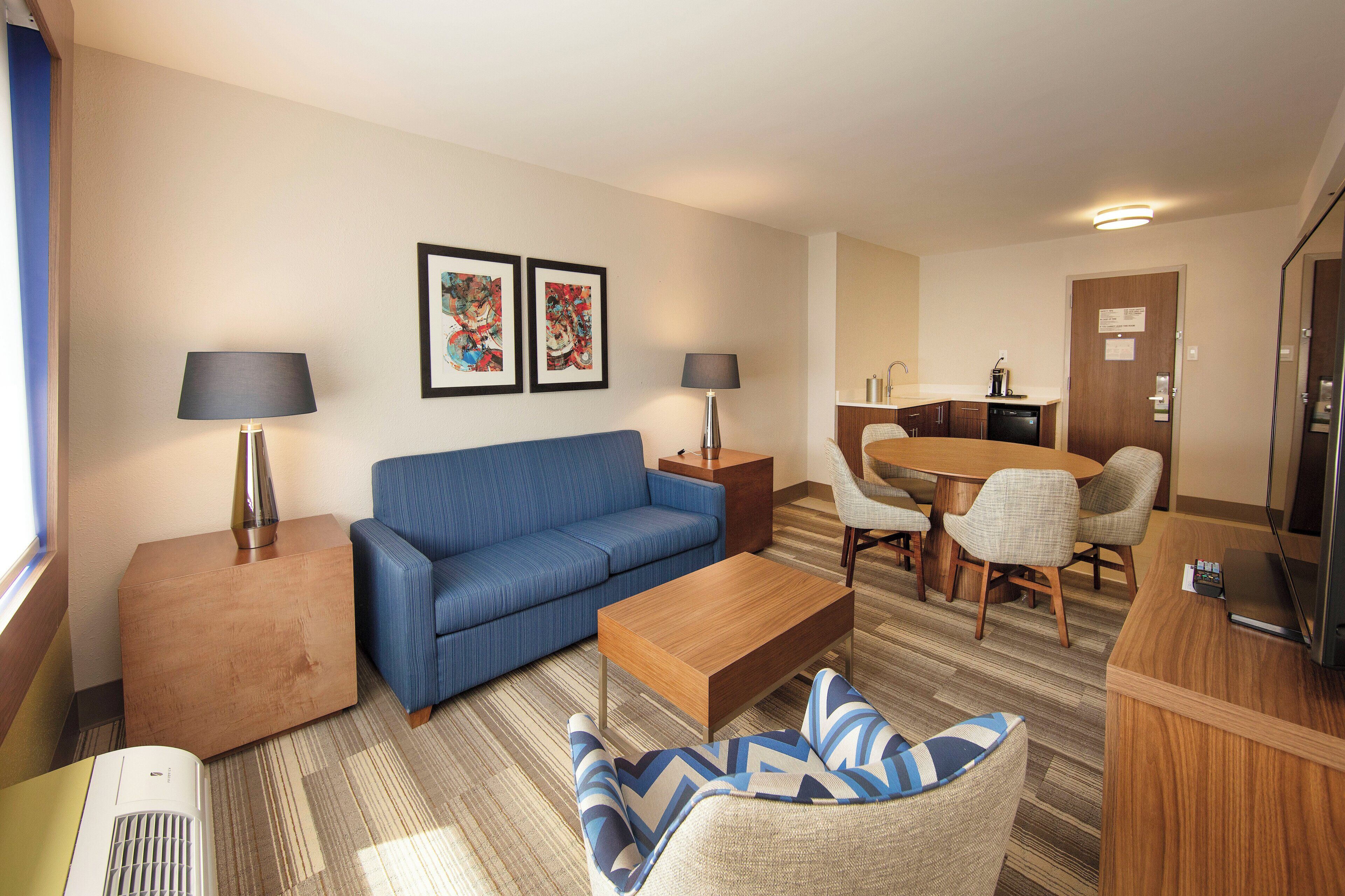 A hotel room with a blue sofa, a wooden coffee table, a patterned rug, a dining table with chairs, and a kitchenette area.