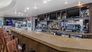 A bar area with a granite countertop, wooden chairs, a TV mounted on the wall, and shelves stocked with bottles and glasses. - Best Western Canoga Park Motor Inn (Winnetka)