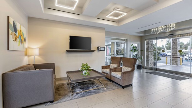 A modern lobby with a sofa, armchair, and a coffee table with a plant. A flat-screen TV is mounted on the wall, and there is a large window with a view of the parking lot. - Best Western Canoga Park Motor Inn (Winnetka)