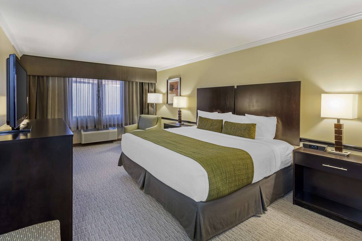 A hotel room with a large bed, two bedside tables with lamps, a desk with a chair, a flat-screen TV, and a large window with curtains.