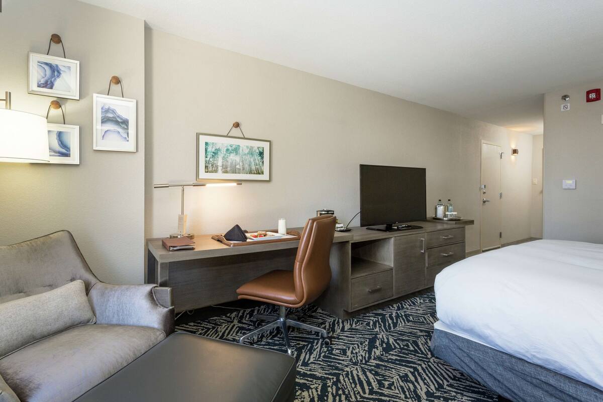 A hotel room with a bed, a desk with a chair, a television, and artwork on the wall.
