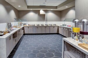Un área de desayuno moderna con piso de baldosas hexagonales, una mesa de buffet con dispensadores de alimentos y una estación de zumos.