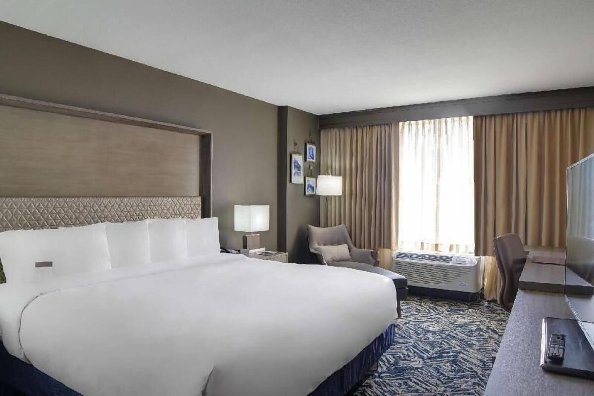 A hotel room with a large bed, a desk, a chair, a TV, and a window with curtains.
