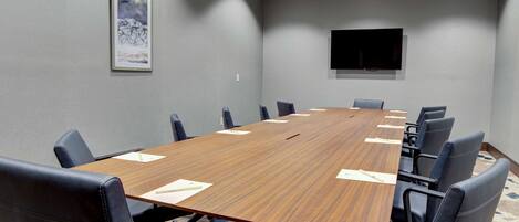 Una sala de conferencias con una larga mesa de madera, sillas de cuero negro, un televisor de pantalla plana montado y una obra de arte enmarcada en la pared.