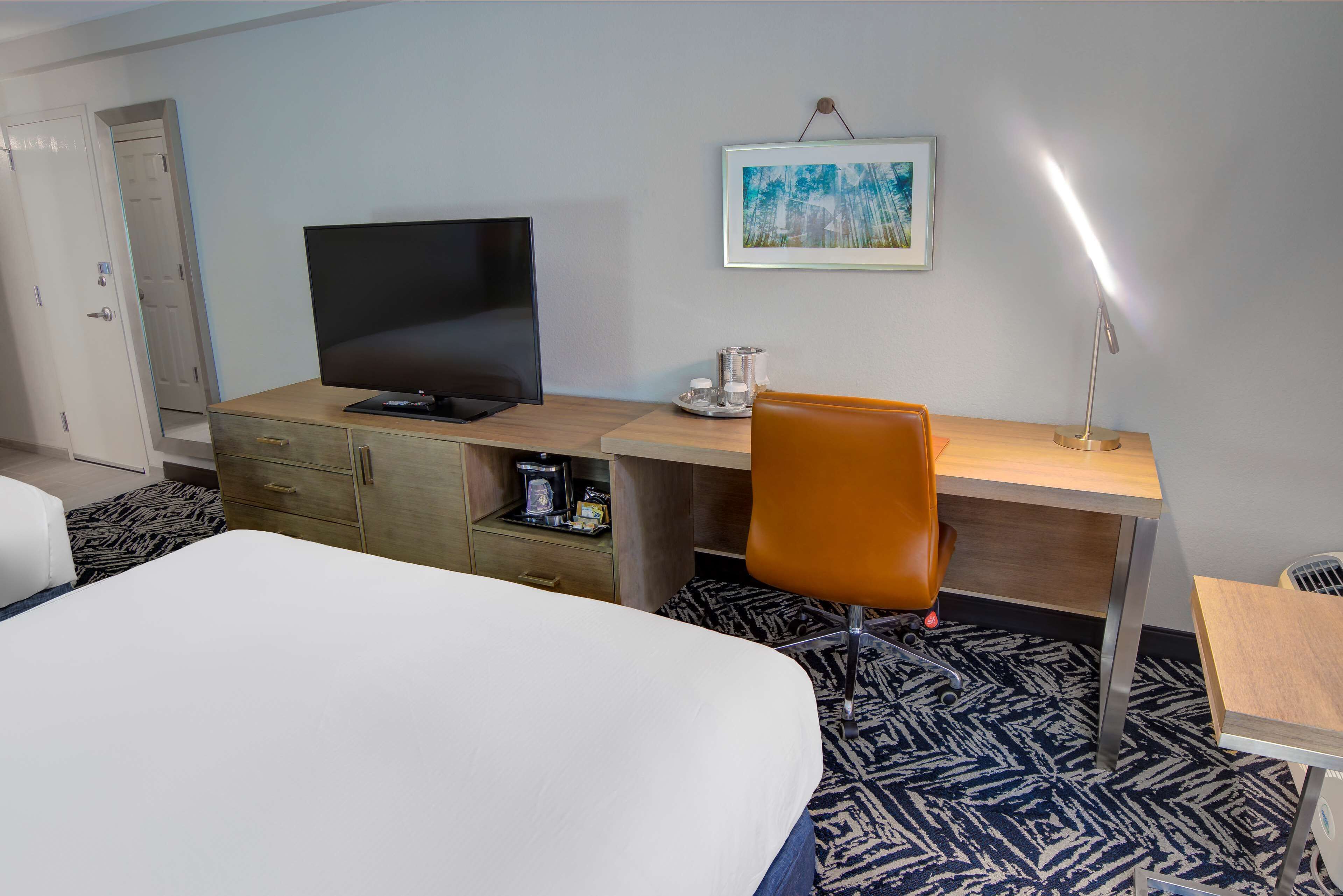 A hotel room with a bed, a desk with a television, a chair, a lamp, and a painting on the wall.