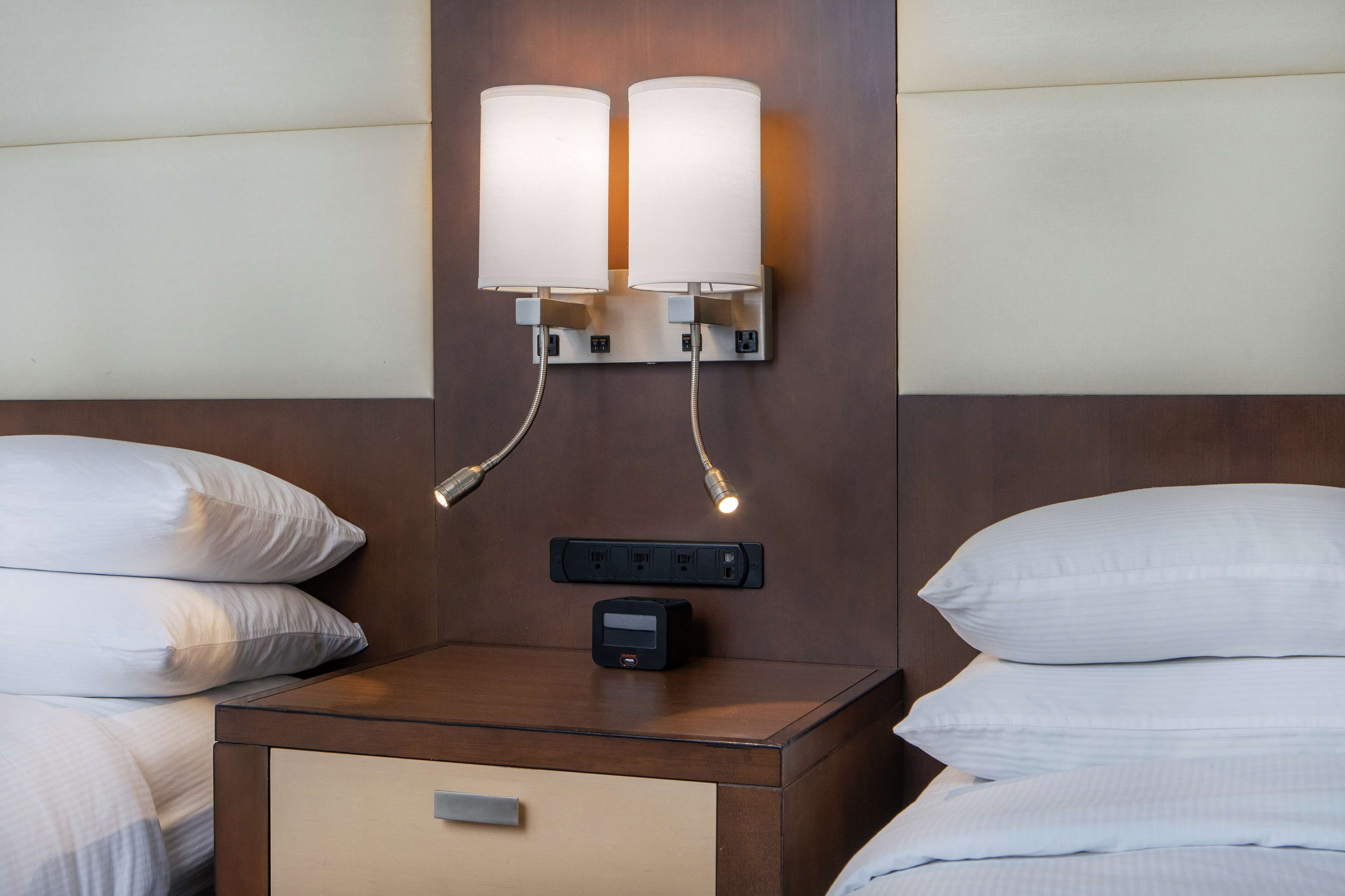 Habitación de hotel con cabecera de madera, dos lámparas de noche, una mesita de noche con un reloj digital y una cama con ropa blanca.