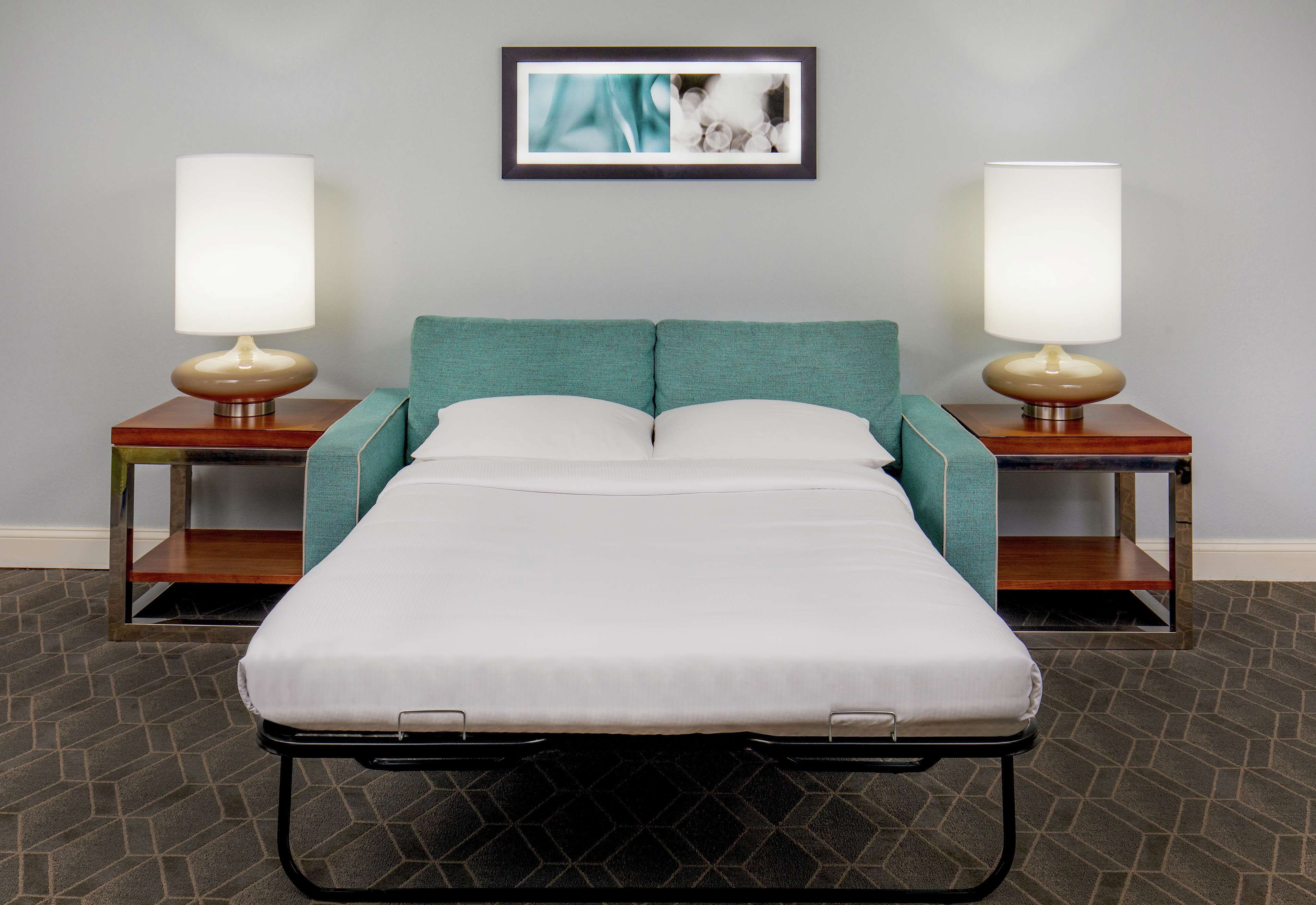 Una cama individual con cabecera color verde azulado, flanqueada por dos mesitas de noche con lámparas, en una habitación con alfombra de diseño geométrico.