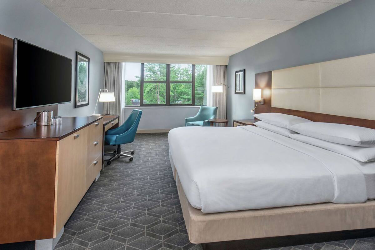 Quarto de hotel com cama, escrivaninha, cadeira, televisão e janela grande.