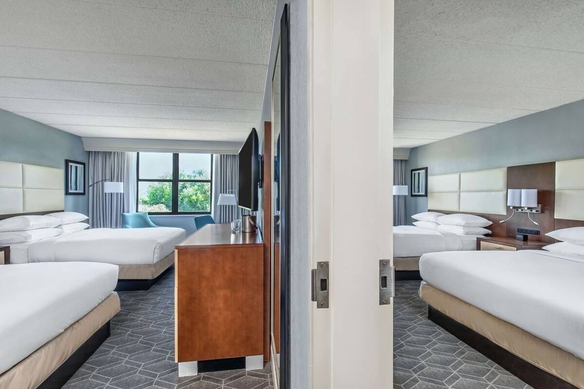 Quarto de hotel com duas camas, uma escrivaninha, uma cadeira, televisão e janela com vista.