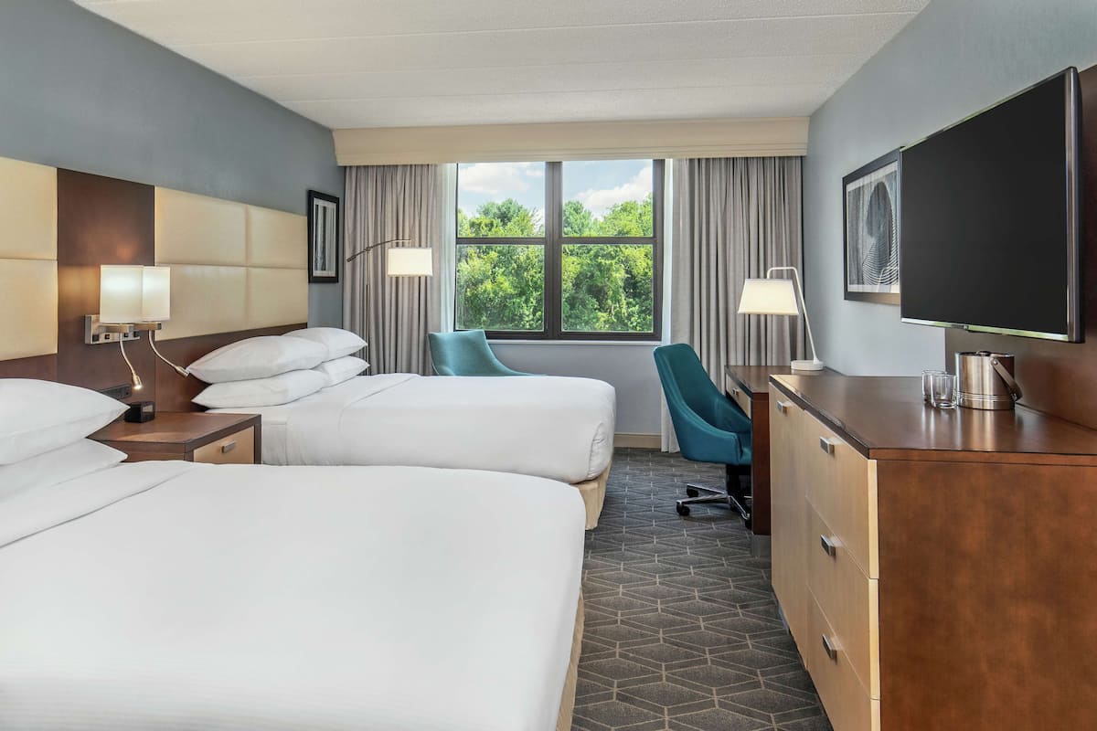Quarto de hotel com duas camas, uma escrivaninha, uma cadeira, televisão e janela com vista para árvores.