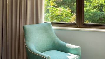 Uma poltrona azul-esverdeada perto de uma janela com cortinas, uma luminária e vista para o verde lá fora.