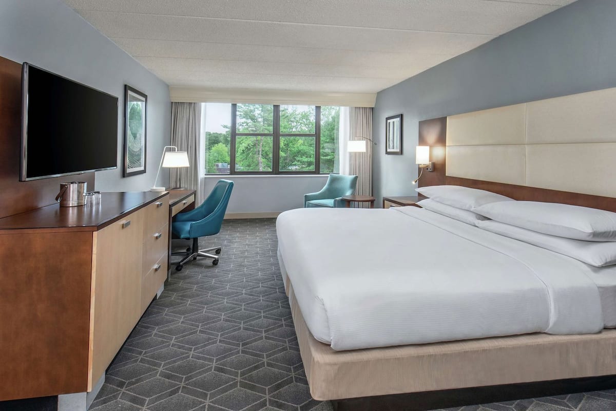 Quarto de hotel com cama, escrivaninha, cadeira, televisão e janela grande.