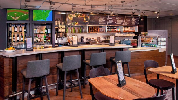 A bar area with a counter, bar stools, and tables. There are shelves with bottles and glasses, and a menu board above the counter.