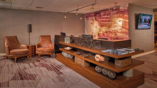 A reception area with two leather chairs, a wooden reception desk, a large abstract painting, and a television screen displaying a news program. - Courtyard by Marriott Wilmington Downtown (Wilmington)