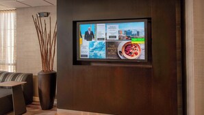 A modern hotel lobby with a large flat-screen TV displaying news and local information, a seating area with a sofa and a coffee table, and a tall vase with branches.