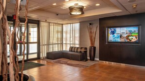 A modern lobby with a seating area, a large screen displaying information, and a decorative plant arrangement.