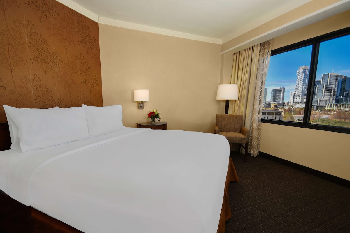 A hotel room with a large bed, two bedside lamps, a chair, a small table with a vase, and a city view through large windows.