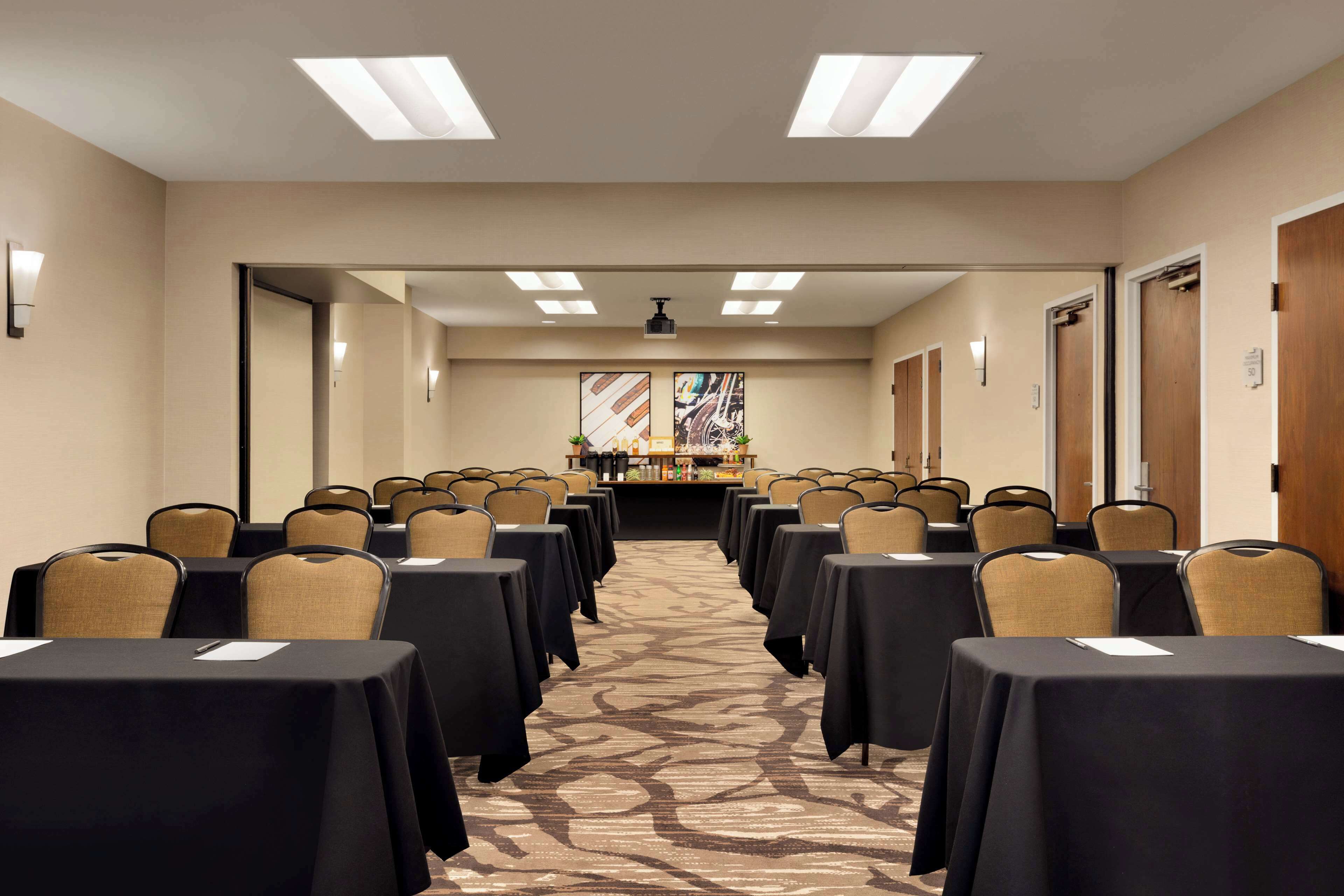 Uma sala de conferências com fileiras de mesas e cadeiras, piso coberto de carpete e um espaço de bar com balcão e cadeiras.