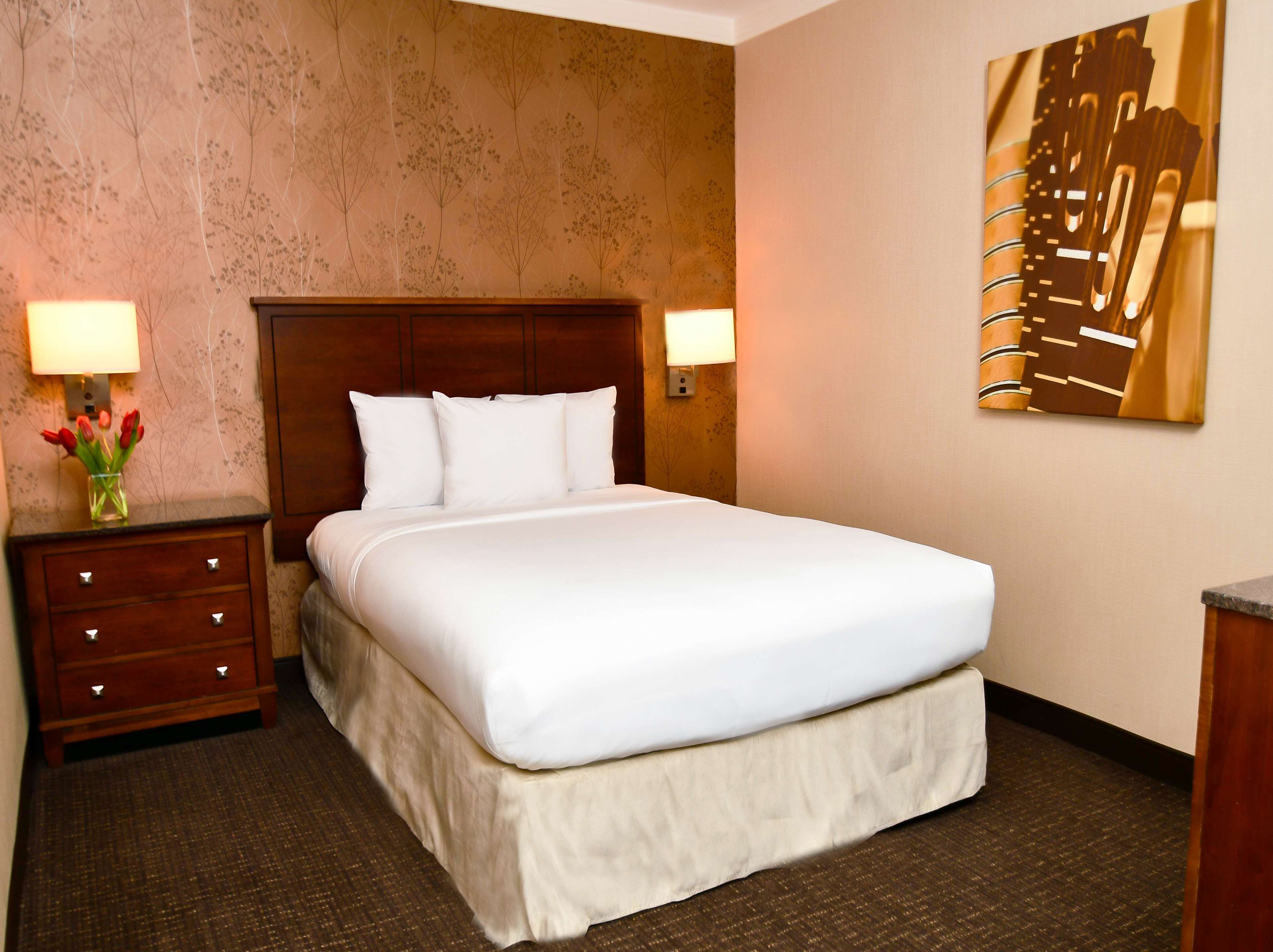 A hotel room with a large bed, two bedside tables with lamps, a vase of red flowers, and a wall-mounted artwork.