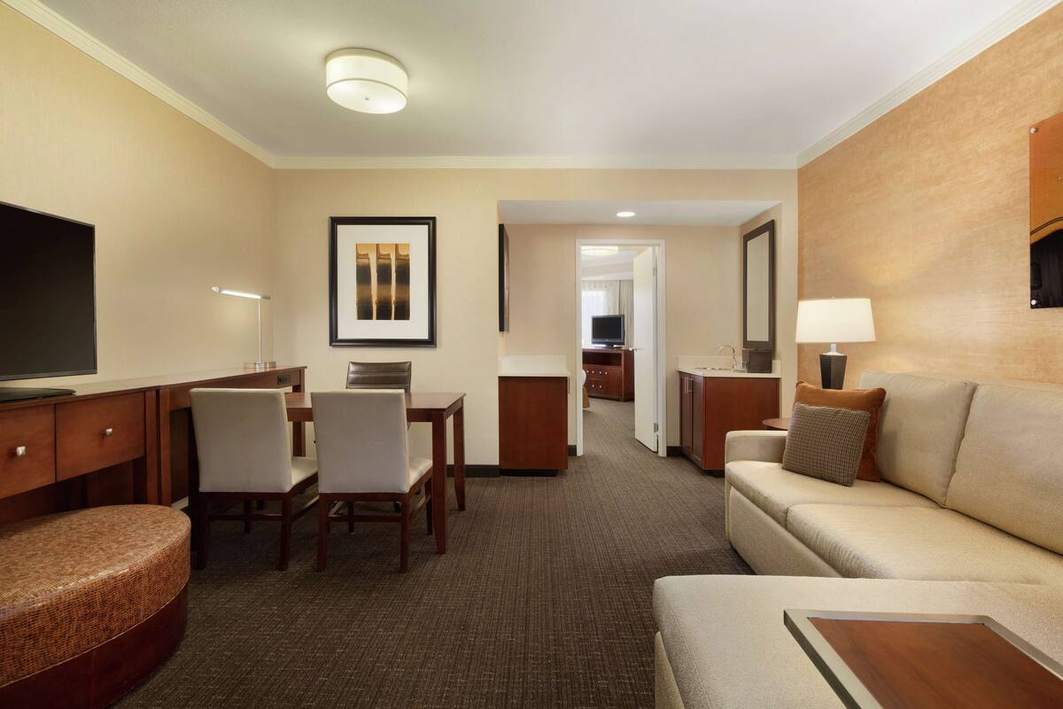 A hotel room with a flat-screen TV, a dining area with chairs and a table, a sofa, a small ottoman, and a lamp.