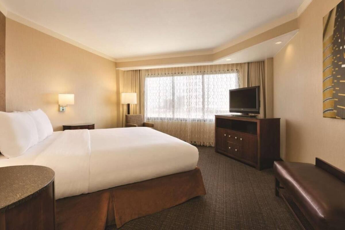 A hotel room with a large bed, a TV, a desk, and a bench.