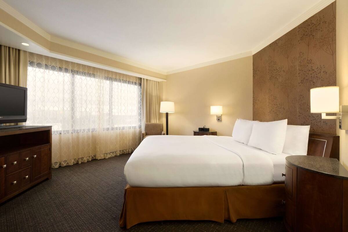 A hotel room with a large bed, a TV on a wooden cabinet, a desk with a lamp, and a wall-mounted light.
