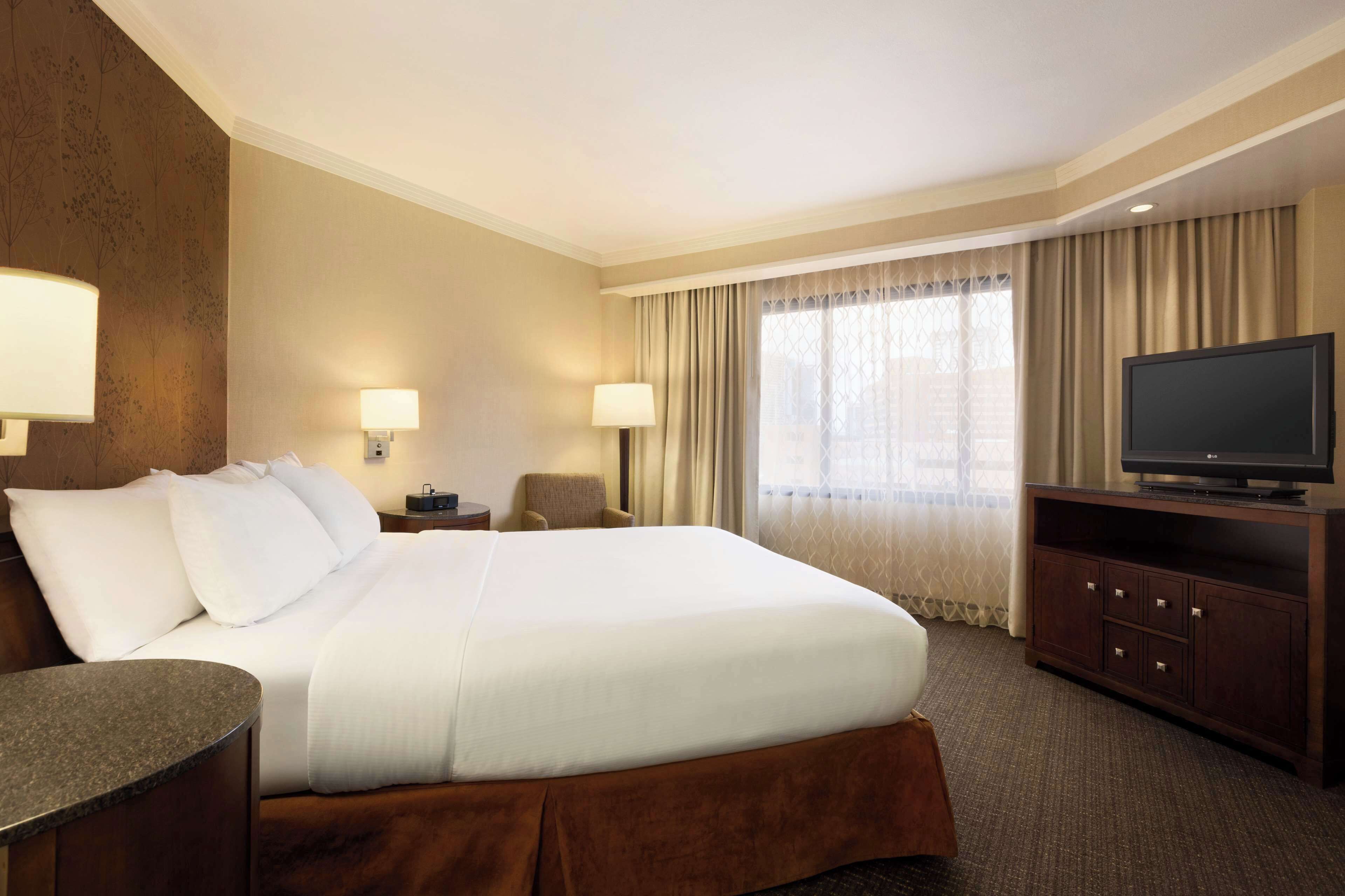 A hotel room with a large bed, two bedside lamps, a television on a wooden cabinet, and a window with curtains.