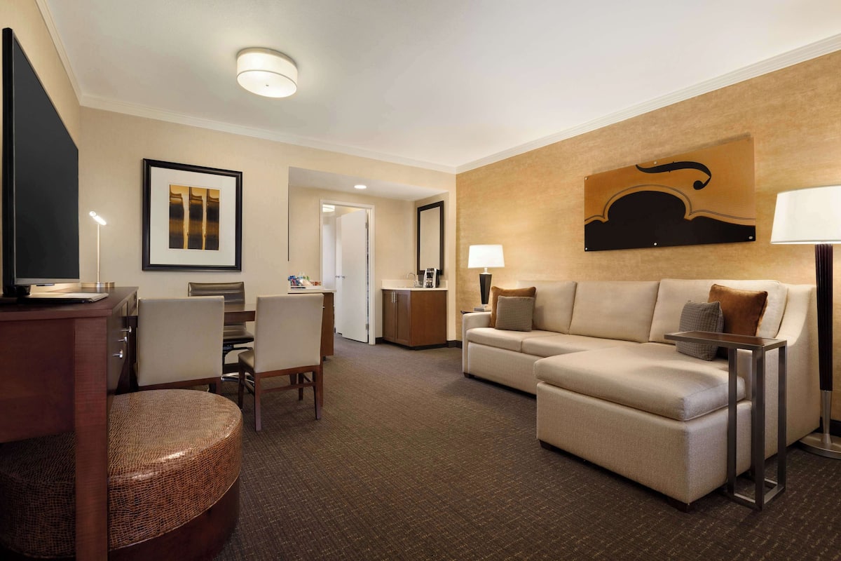 A hotel room with a living area featuring a sofa, a dining table with chairs, a TV, and a lamp.