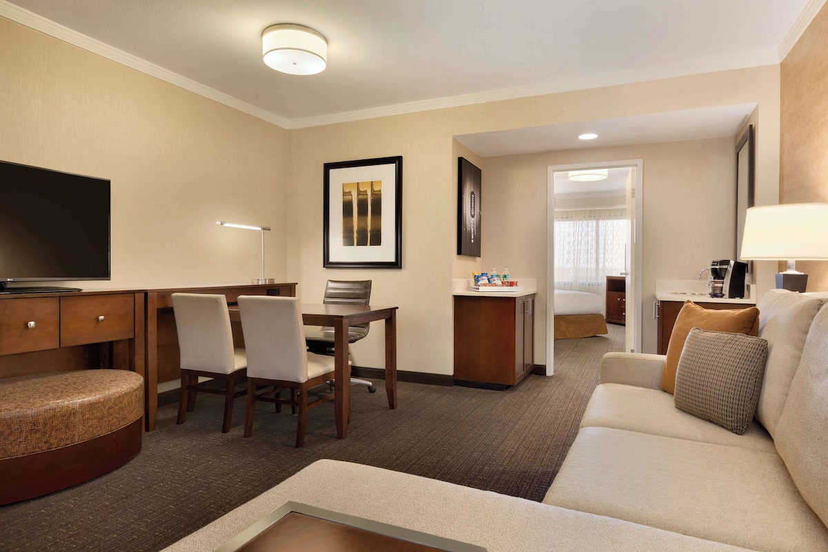 A hotel room with a sofa, a TV, a desk with chairs, a small kitchen area, and a bathroom visible through an open door.