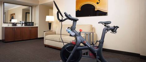 A Peloton exercise bike in a hotel room with a large mirror, a sofa, and a painting on the wall.