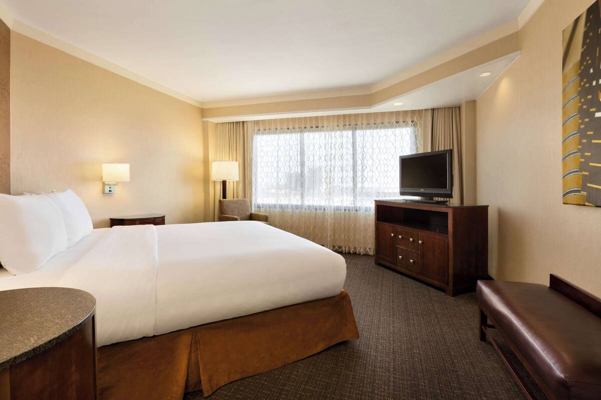A hotel room with a large bed, a television, a desk, and a bench.