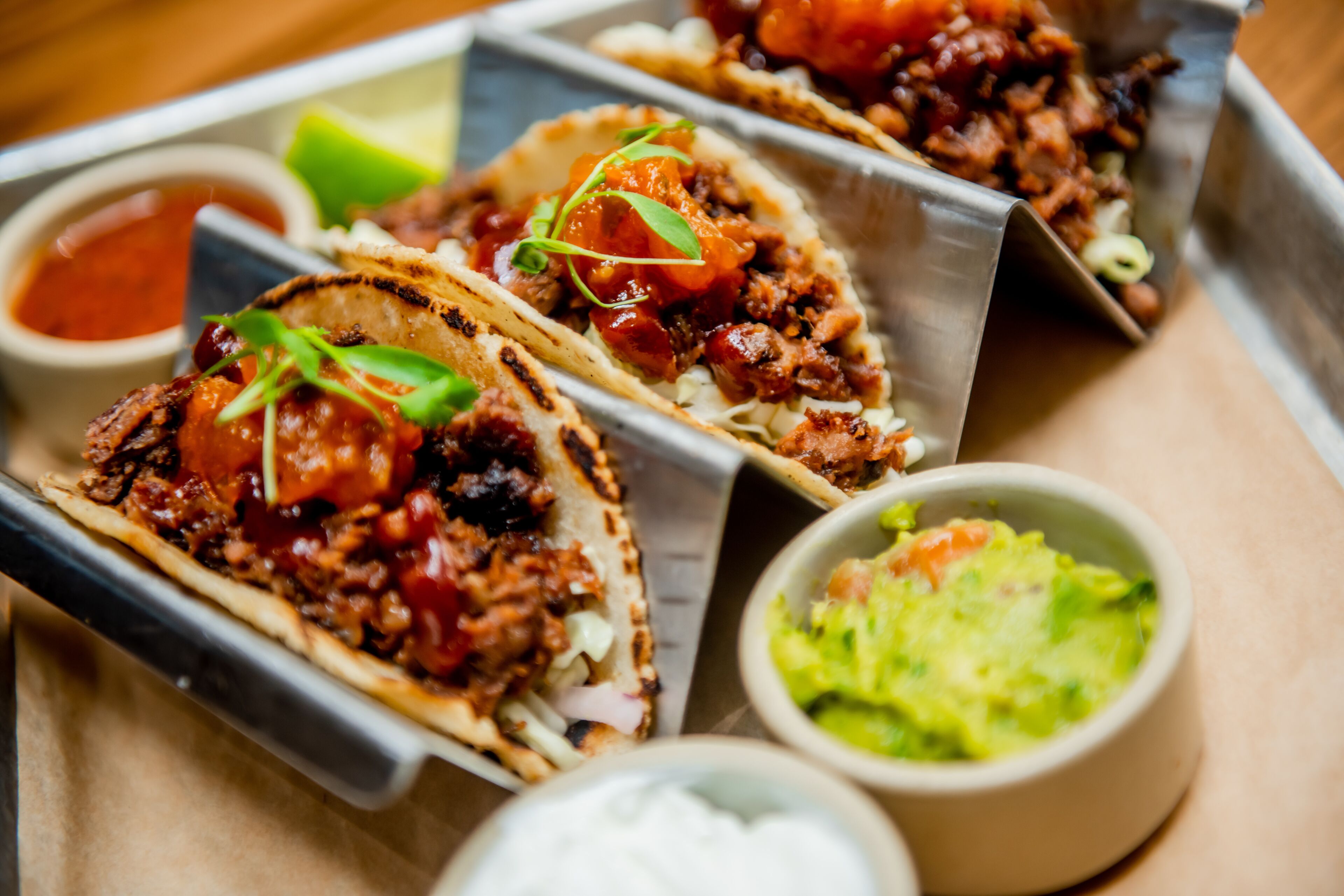 Three tacos with various toppings and sides including a bowl of guacamole and a small bowl of sauce.