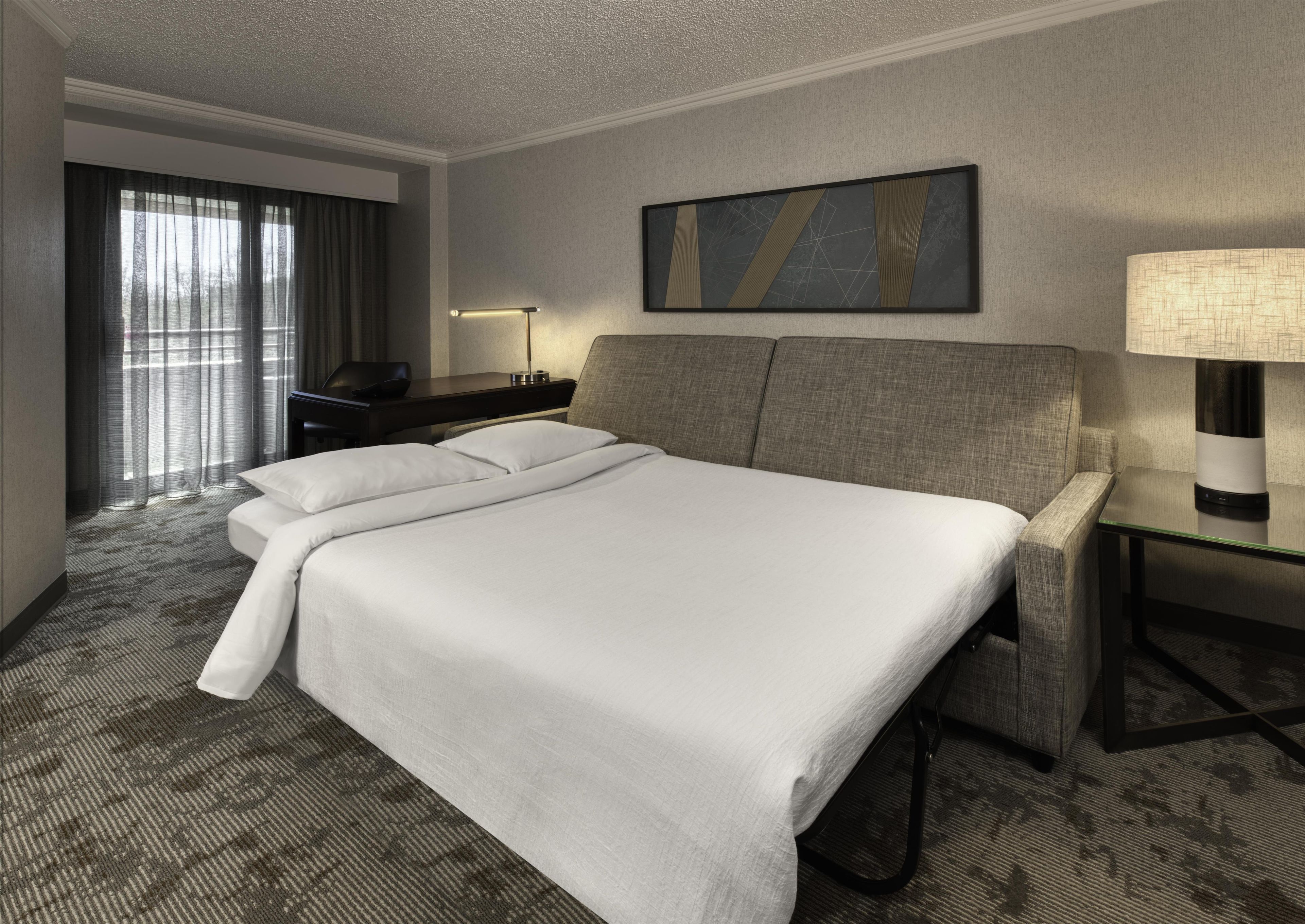 Una habitación de hotel con una cama grande, un escritorio, una silla, una lámpara y un cuadro en la pared.