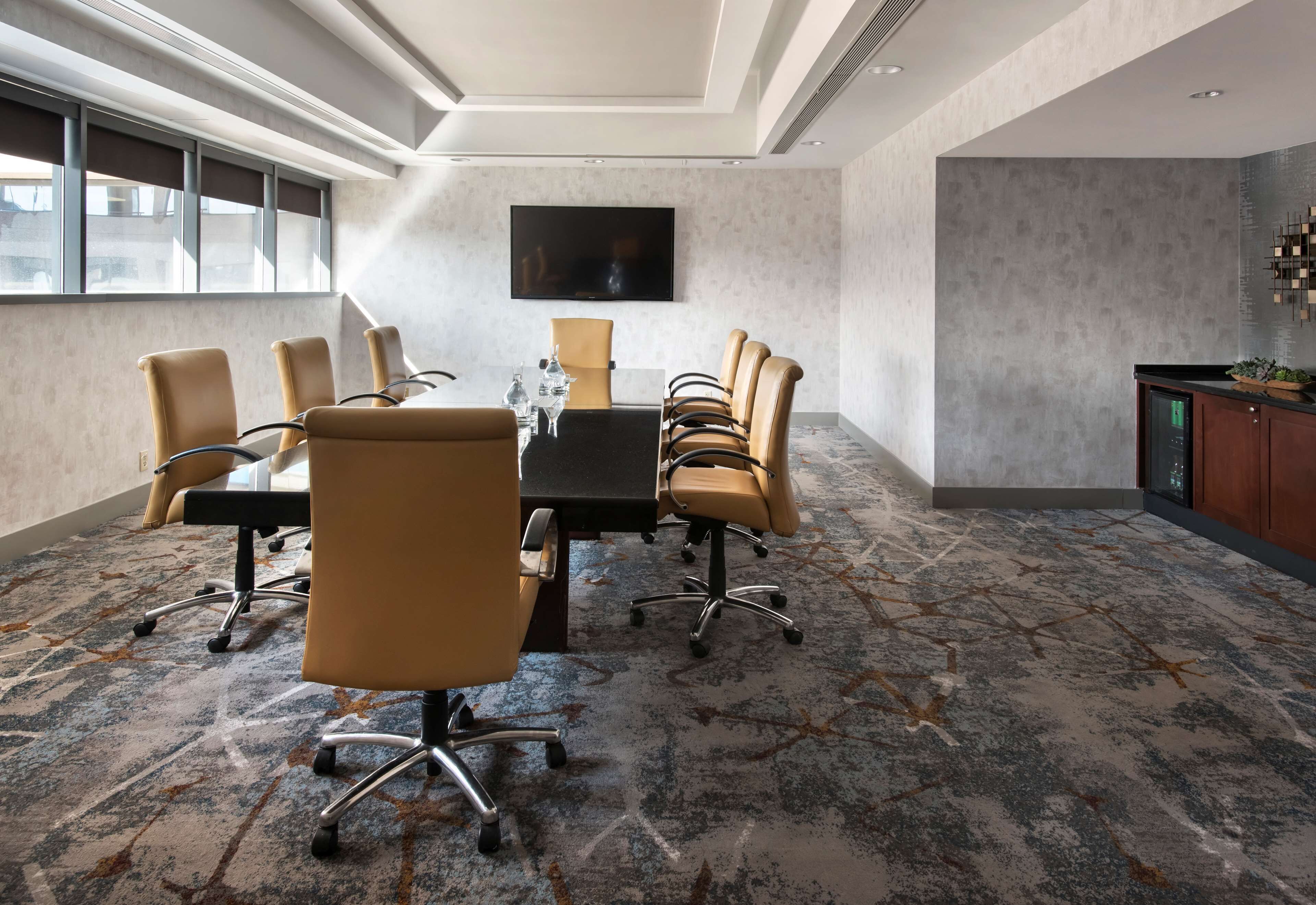 A conference room with a large table, multiple chairs, a flat-screen TV, and a patterned carpet.