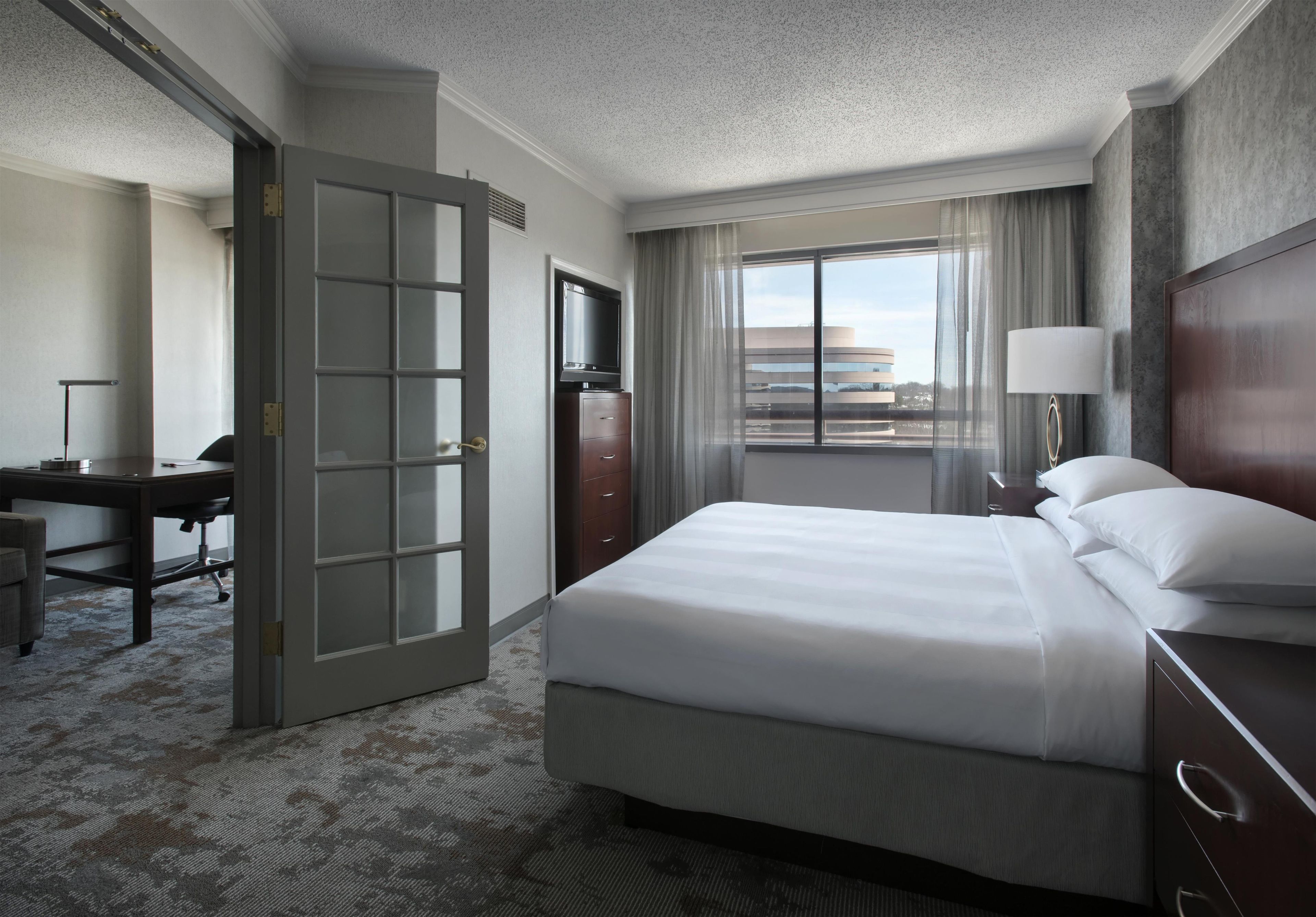 Une chambre d’hôtel comprenant un lit, un bureau, une chaise, une commode, un téléviseur, une fenêtre avec des rideaux et une lampe.