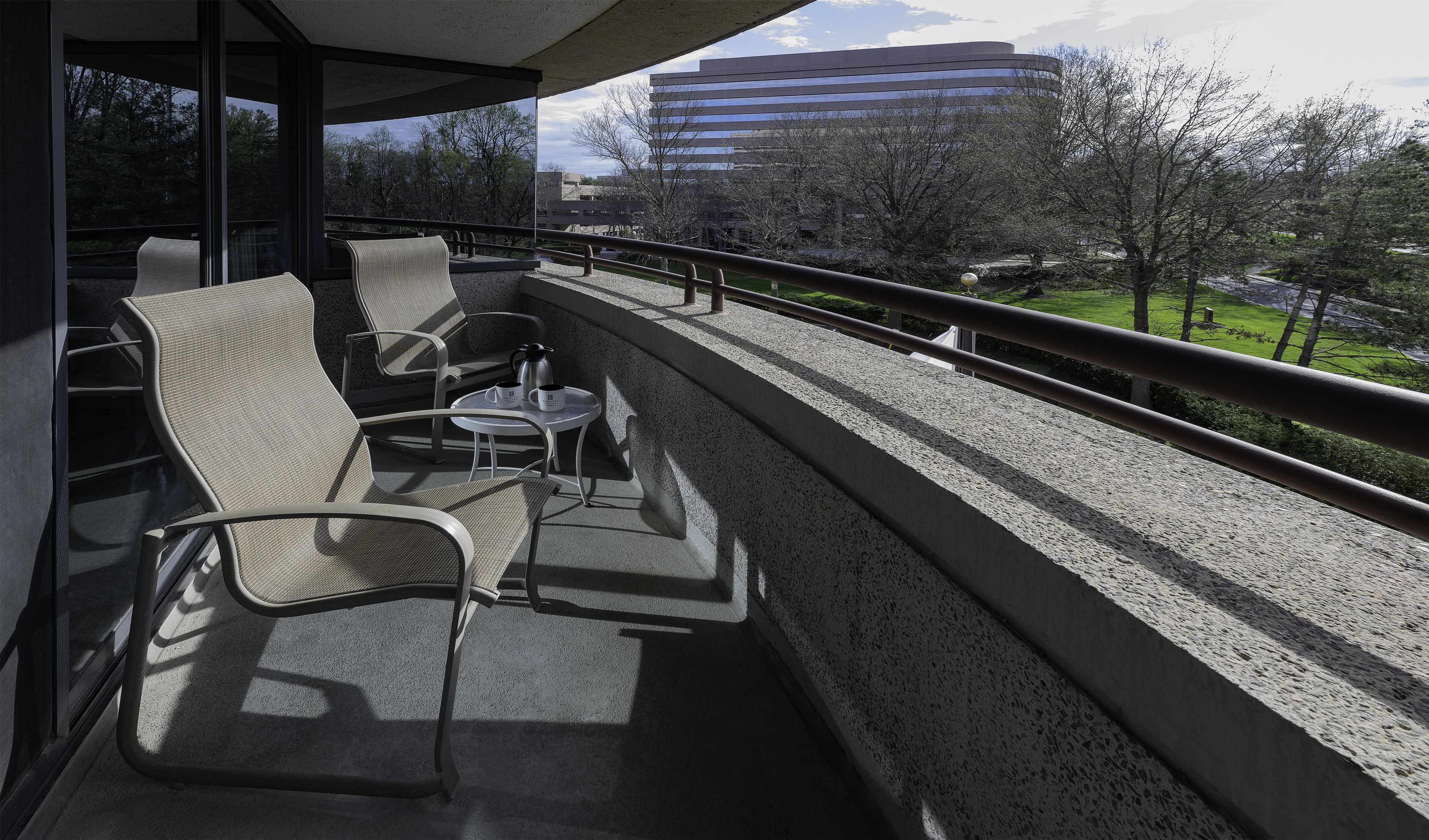 Un balcón con mobiliario exterior, una mesa y vistas a un edificio y árboles.