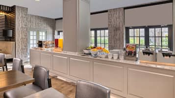 Un bar à petit-déjeuner moderne proposant une variété de fruits et de boissons, un espace détente avec des tables et des chaises, et de grandes fenêtres offrant une vue sur l’extérieur.